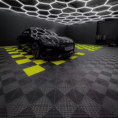 Ein schwarzes Auto steht auf einem modernen, gestreiften Bodenbelag, der aus dunkelgrauen und leuchtend gelben Fliesen besteht. Eine hexagonale Beleuchtung reflektiert sich auf der Karosserie des Autos in einem dunklen Raum.